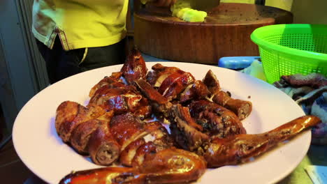 Primer-Plano-De-Una-Carne-De-Pato-Con-2-Cabezas-Colocadas-En-Un-Plato-Con-Las-Manos-De-Un-Hombre-Cortando-Y-Sosteniendo-Un-Cuchillo-Grande-En-El-Fondo