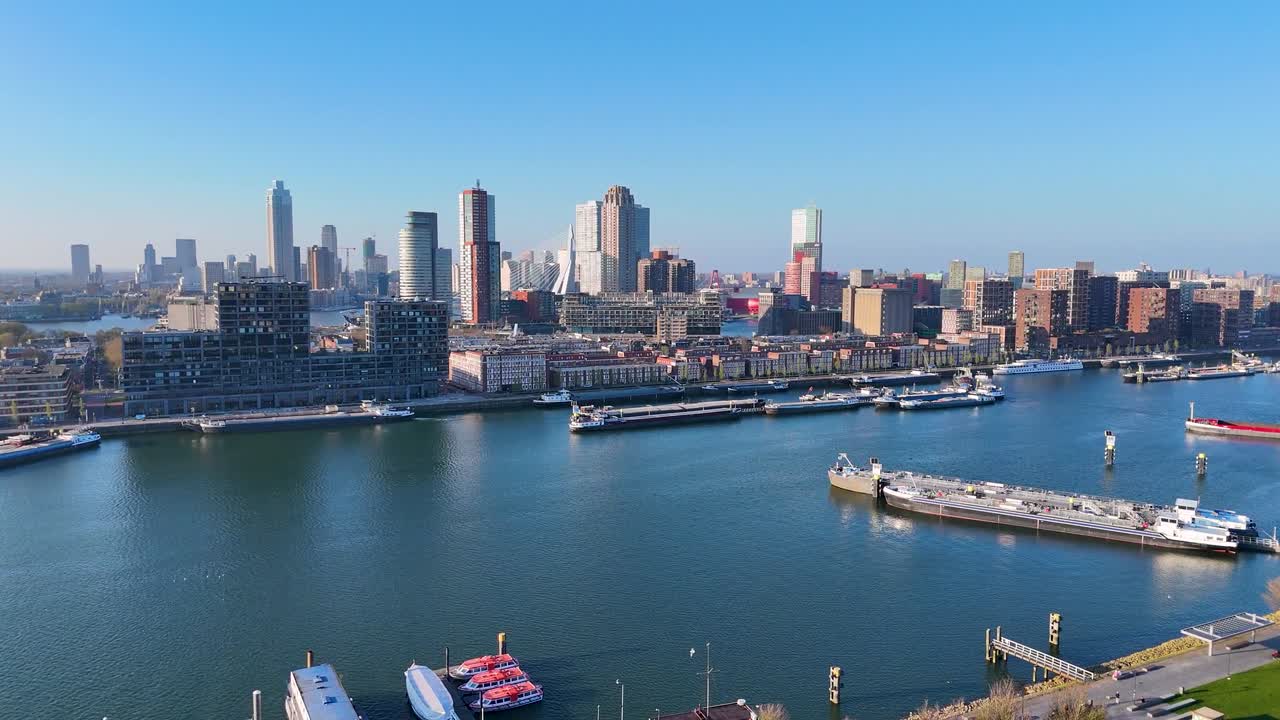 Aerial drone footage of waterfront skyline and active harbor in Kop van Zuid Rotterdam Netherlands showing modern towers cargo ships and cityscape under bright clear blue sky