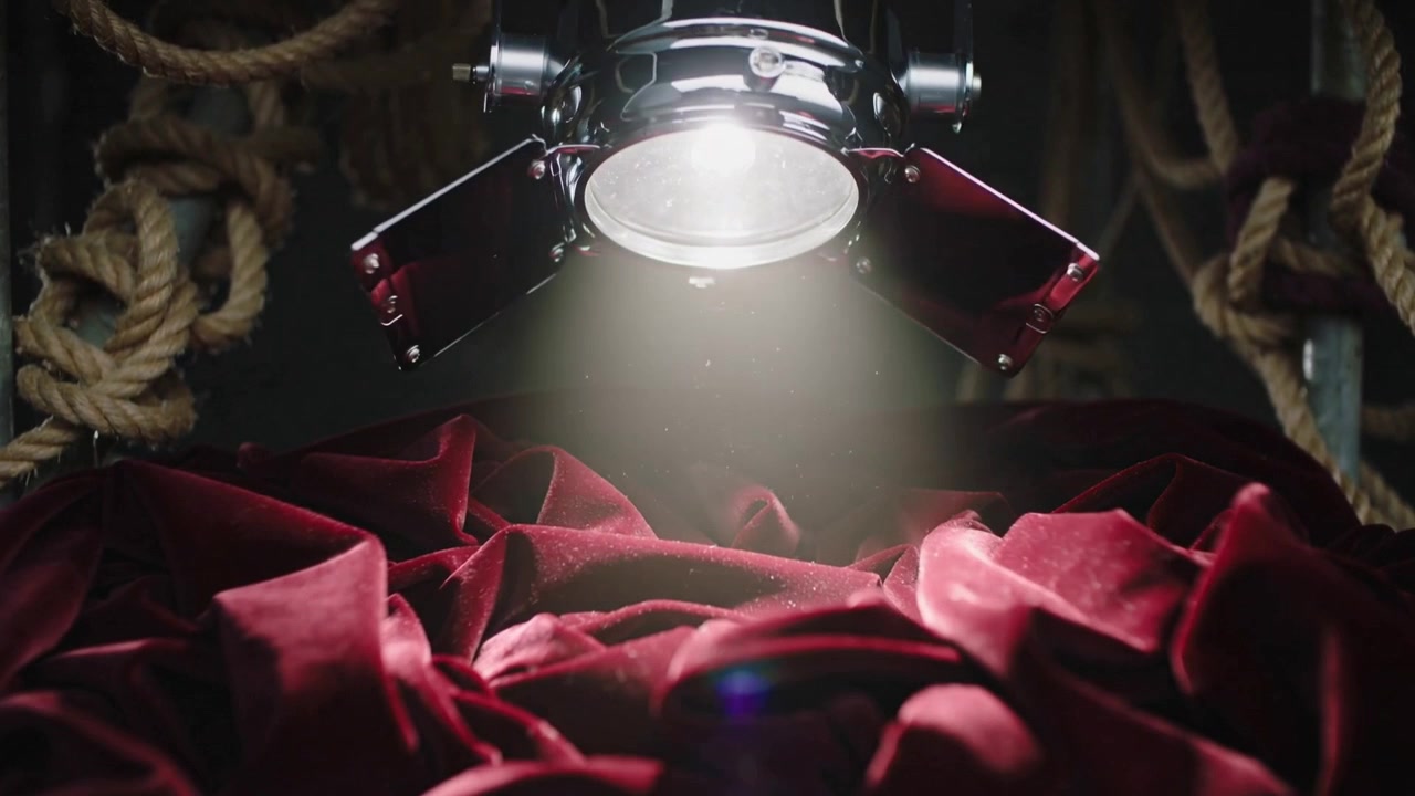 Faro cromato che si abbassa, rivelando profonde pieghe di velluto rosso sul palco per una presentazione, con corde arrotolate.