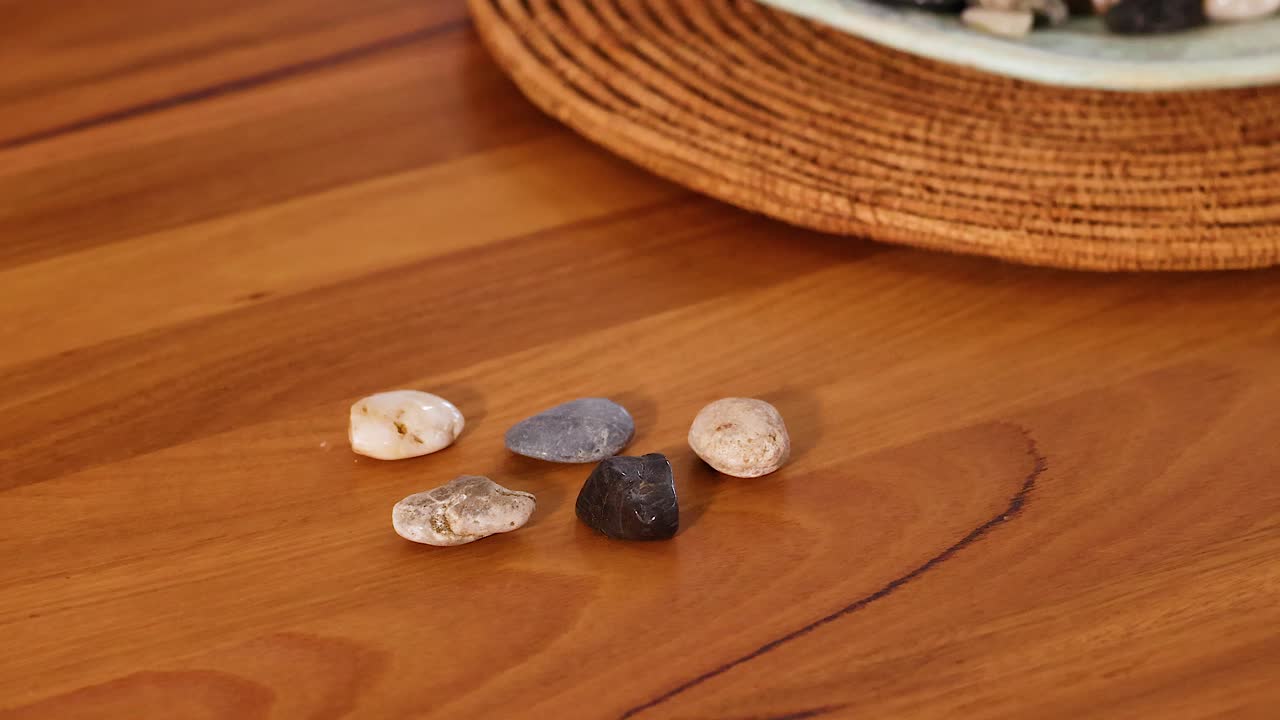 A hand places a black stone among others on a wooden table, creating a balanced arrangement in warm lighting