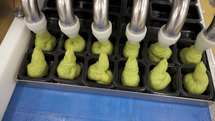 Stainless depositor tubes squeeze green avo HPP mix into neat mounds inside black plastic trays on a moving belt.