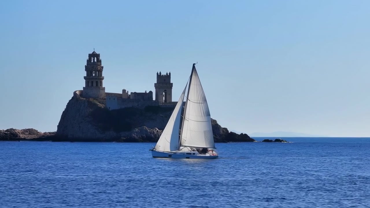Uno yacht a vela si muove costantemente sull'acqua leggermente increspata mentre le vele bianche illuminate dal sole si stagliano contro il mare blu profondo e il cielo pallido, con rocce scoscese e storiche torri in pietra sullo sfondo