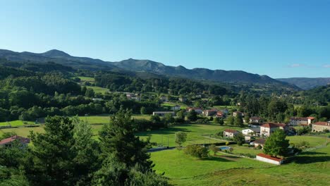 vuelo de avión no tripulado en ascenso mirando las copas de los árboles en el área vasco-cantabria sobre una ciudad rural con tierras de cultivo y exuberantes bosques de roble y montañas en una tarde con cielo azul en verano y hierba verde