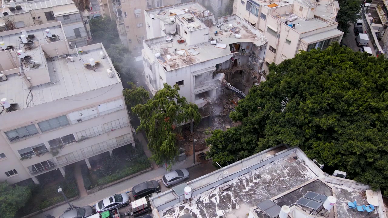 Un drone avanza in alto, rivelando un escavatore che smantella un edificio danneggiato sottostante.