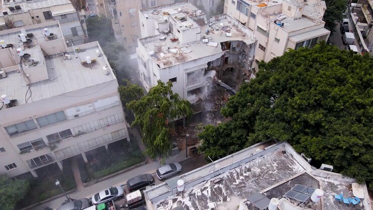 Un drone avanza in alto, rivelando un escavatore che smantella un edificio danneggiato sottostante.