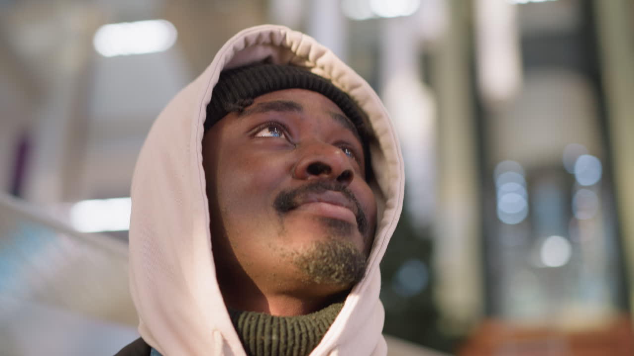 A close-up shot of a young man with a light hoodie and black knit beanie, looking upwards with a smile. His facial features are expressive. The blurred background features bright lights, a shopping mall