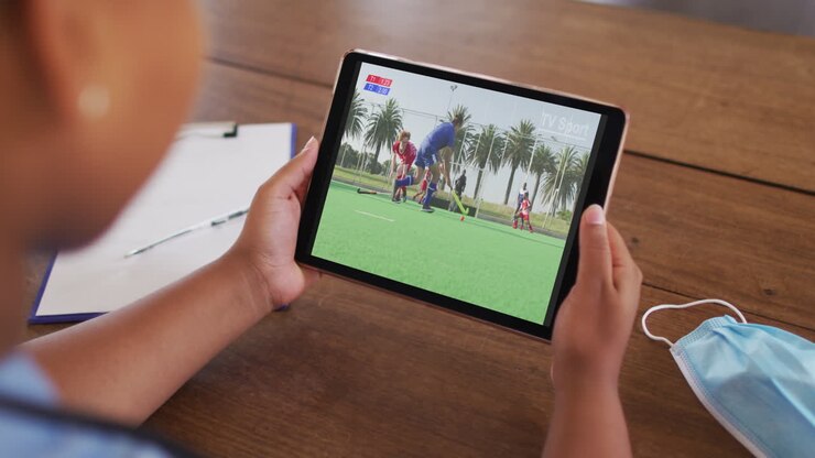 Composite of woman sitting at table with face mask, watching hockey match on tablet