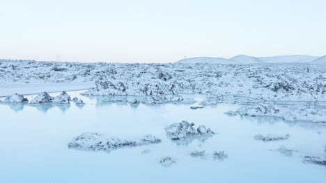 Fliegen-über-Lebendiges-Blaues-Geothermisches-Wasser-In-Einem-Felsigen-Lavafeld,-Das-Mit-Schnee-Bedeckt-Ist