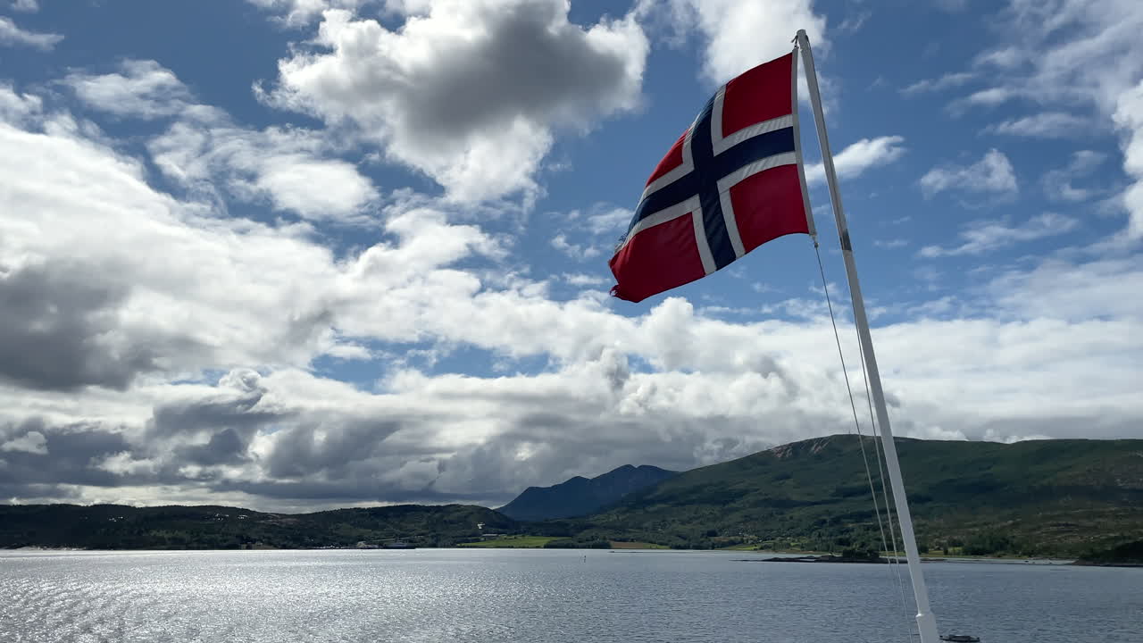 Norsk flagg som vaier i vinden fra et fergedekk med en naturskjønn bakgrunn på en solfylt dag
