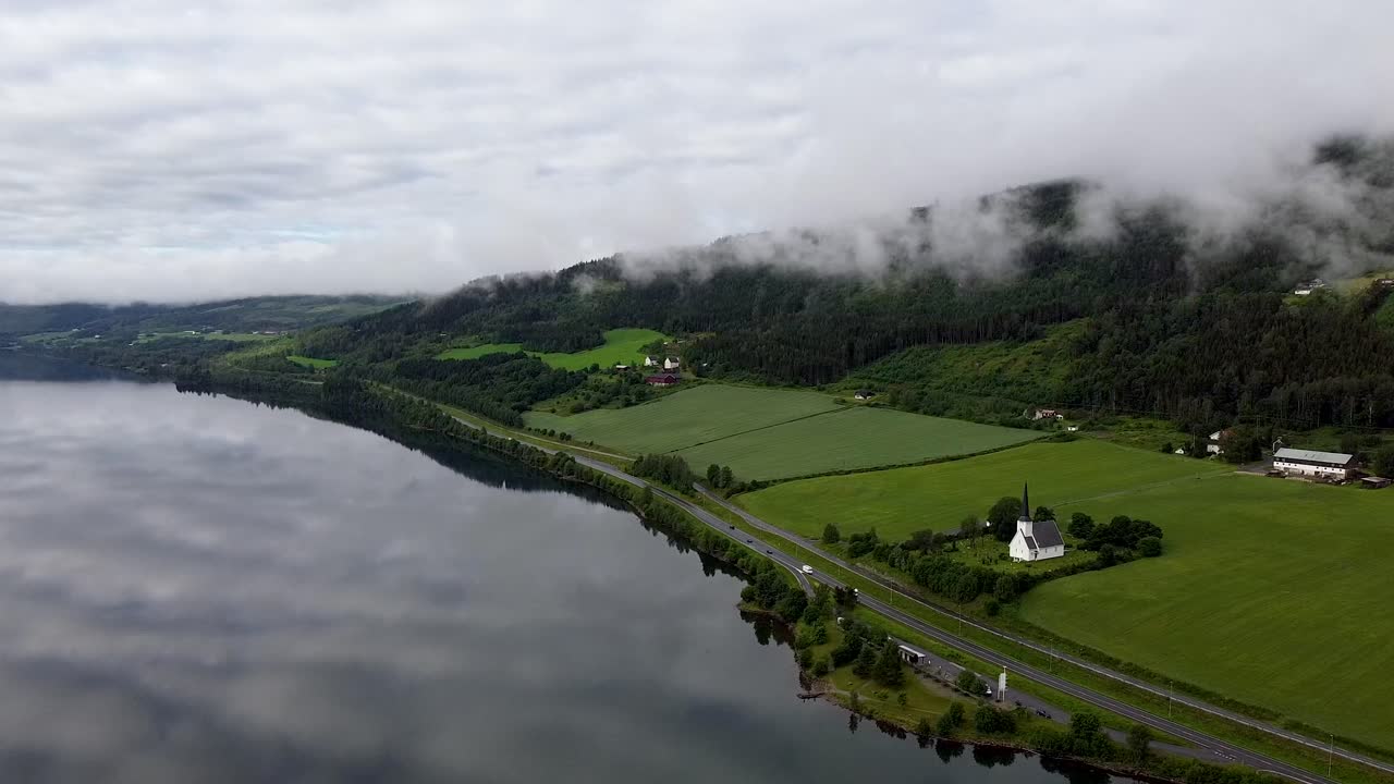 imágenes aéreas que muestran la iglesia de vingrom, el lago mjøsa y la carretera cercana con un telón de fondo de campos verdes y colinas cubiertas de niebla en noruega