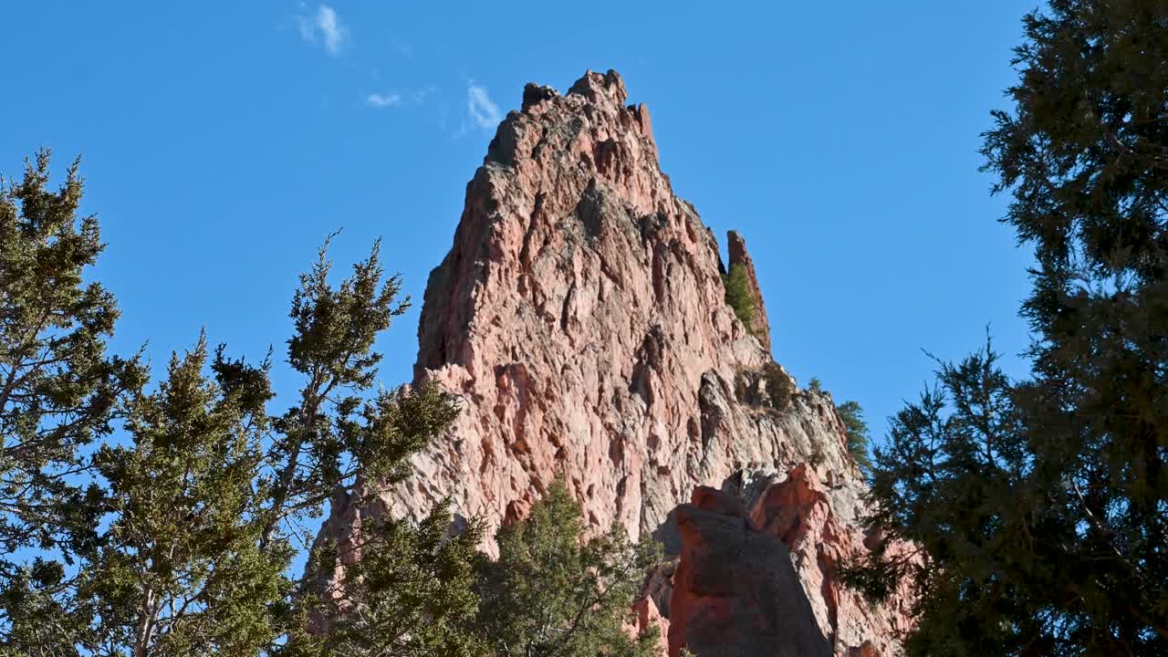 Drone shot of jagged red sandstone pinnacle at Garden of the Gods, framed by treetops and vivid blue sky in Colorado Springs