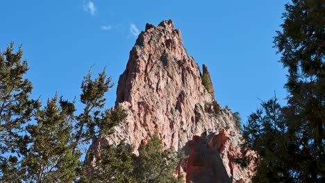 drone shot of jagged red sandstone pinnacle at garden of the gods, framed by treetops and vivid blue sky in colorado springs