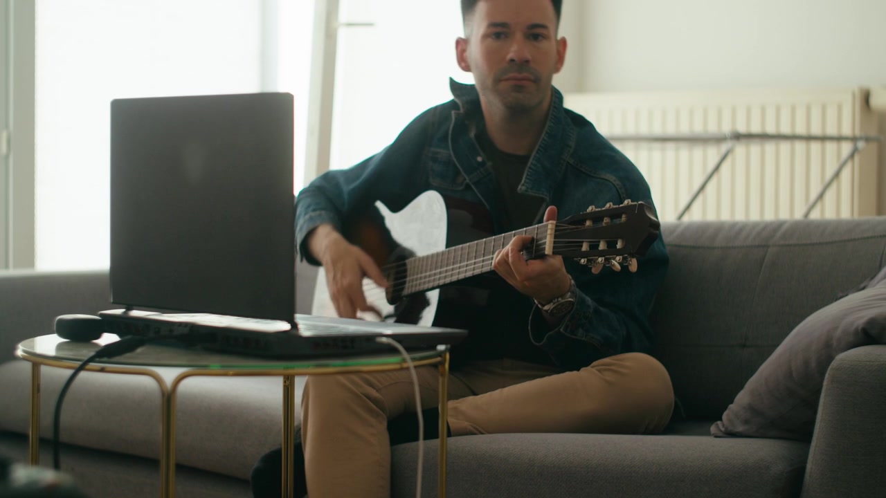 Uzaktan eğitim veya dijital müzik dersleri için dizüstü bilgisayar kullanarak evde akustik gitar pratik yapan genç bir adamın otantik bir sahnesi