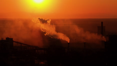 Industrial-smokestacks-with-smoke-and-steam-emissions,-against-the-vivid-hues-of-orange-sunlight,-high-contrast