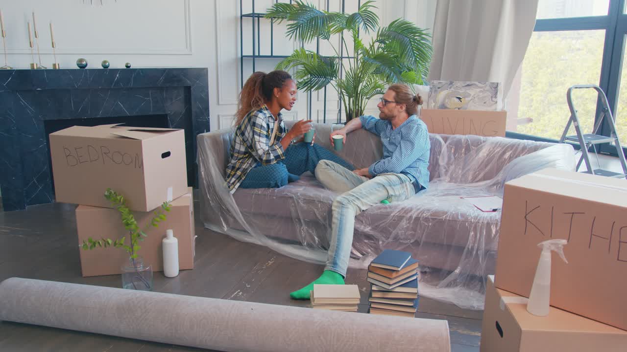 다양한 커플이 아파트로 이사하고 커피를 마시며 상자 사이에 소파에 앉아 있습니다.