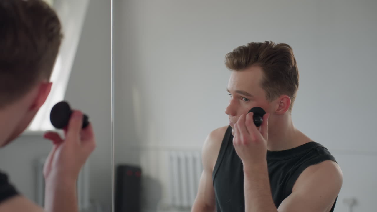 rear view of fair skinned boy applying makeup with sponge before large mirror in bright minimalist studio space with soft daylight illuminating neutral interior and urban reflection