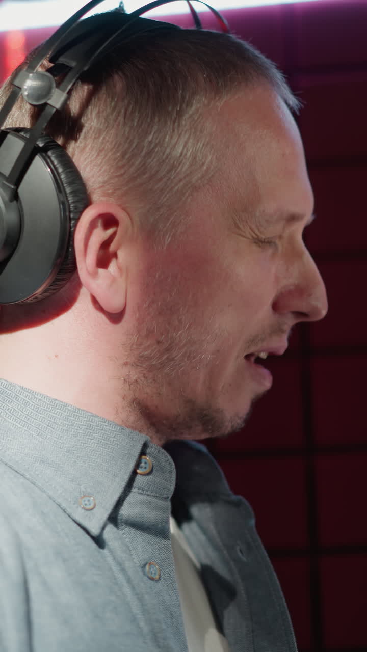 un primer plano de un hombre en un estudio de grabación con una camisa de manga larga azul y auriculares negros, cantando apasionadamente con los ojos cerrados frente a un micrófono, con un fondo de panel acústico rojo