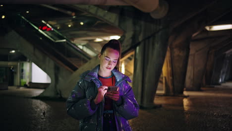 chica glamurosa enviando mensajes de texto teléfono inteligente de pie bajo el puente urbano. mujer surfing