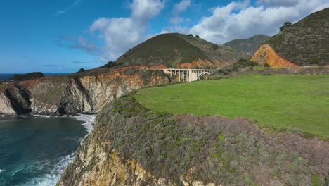 Aerial-view-of-sea-cliffs-revealing-Bixby-bridge-behind-green-fields,-Highway-1-California