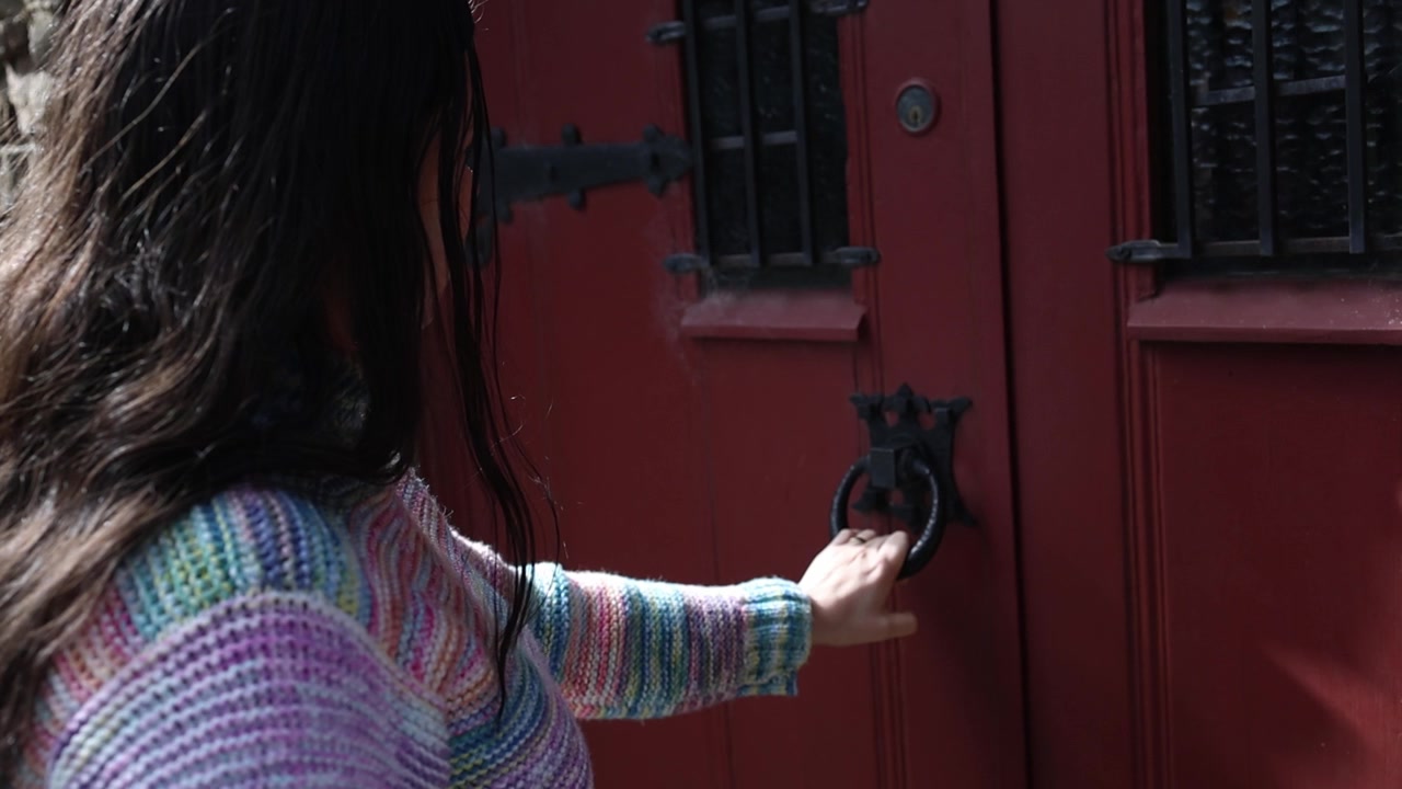 Una donna con un maglione colorato bussa delicatamente a una pesante porta rossa di chiesa, la luce del sole illumina rustiche pareti in pietra e dettagli in ferro battuto. Inquadratura ravvicinata lenta e intima, telecamera stabile