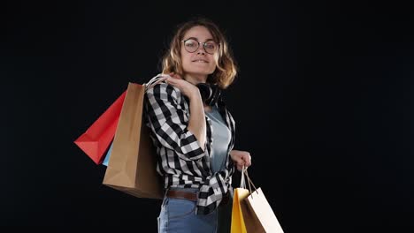 sonriente comprador emocionado joven mujer moderna con auriculares en el cuello y gafas elegantes posando con coloridas bolsas de papel de compras, sosteniendo en el hombro aislado en fondo de estudio negro