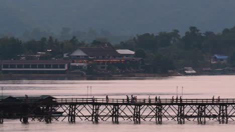 Mon-Bridge-and-a-longboat-followed-speeding-behind-the-structure-while-people-are-walking-on-the-bridge,-silhouetting-as-it-was-getting-dark,-in-slow-motion