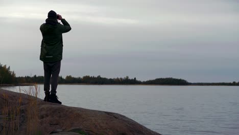 Detrás-Del-Hombre-Parado-En-El-Lago-Mirando-A-Través-De-Binoculares,-Mirando-Al-Lago