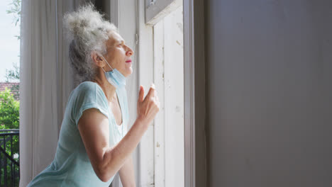 Mujer-Caucásica-Mayor-Con-Mascarilla-Baja-Mirando-Por-La-Ventana-En-Casa