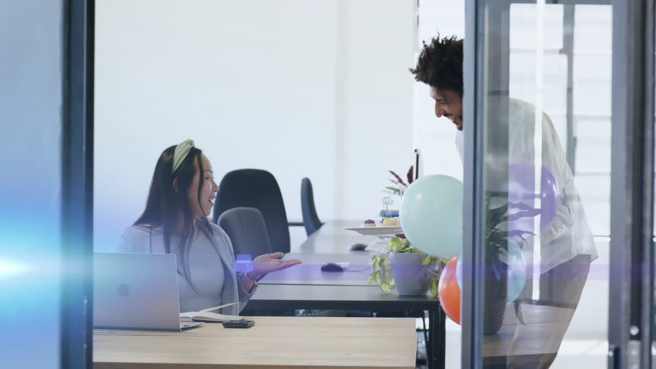 Woman working laptop in office, man appearing via glass with balloons and cake celebrating coworker