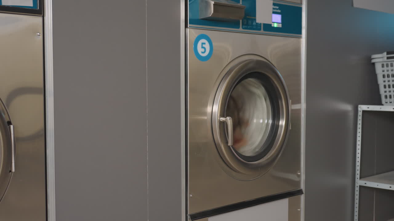 Zoom out on stainless laundry machine labelled 5 showing transparent glass door with clothes spinning inside during wash cycle, reflective surface highlighting motion inside laundromat environment