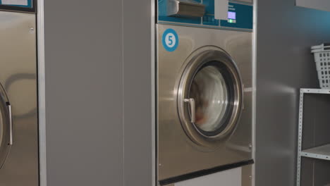 Zoom out on stainless laundry machine labelled 5 showing transparent glass door with clothes spinning inside during wash cycle, reflective surface highlighting motion inside laundromat environment