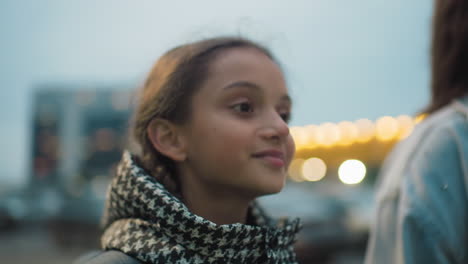 young girl with backpack smiles while walking in evening light near tall apartment buildings, dressed in warm checkered coat, expressing happiness, peace, and curiosity in modern city environment with soft glow