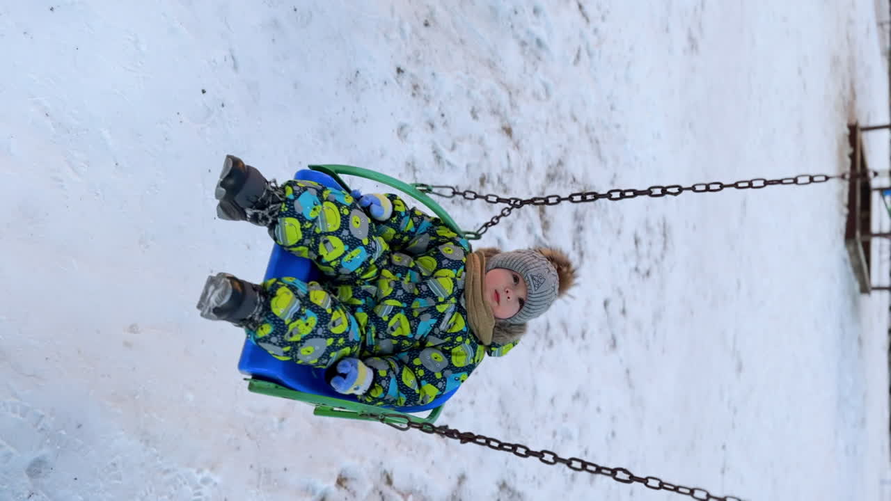 Sweet kid in a warm romper and cap swinging on a swing. Calm baby boy on the walk in winter.