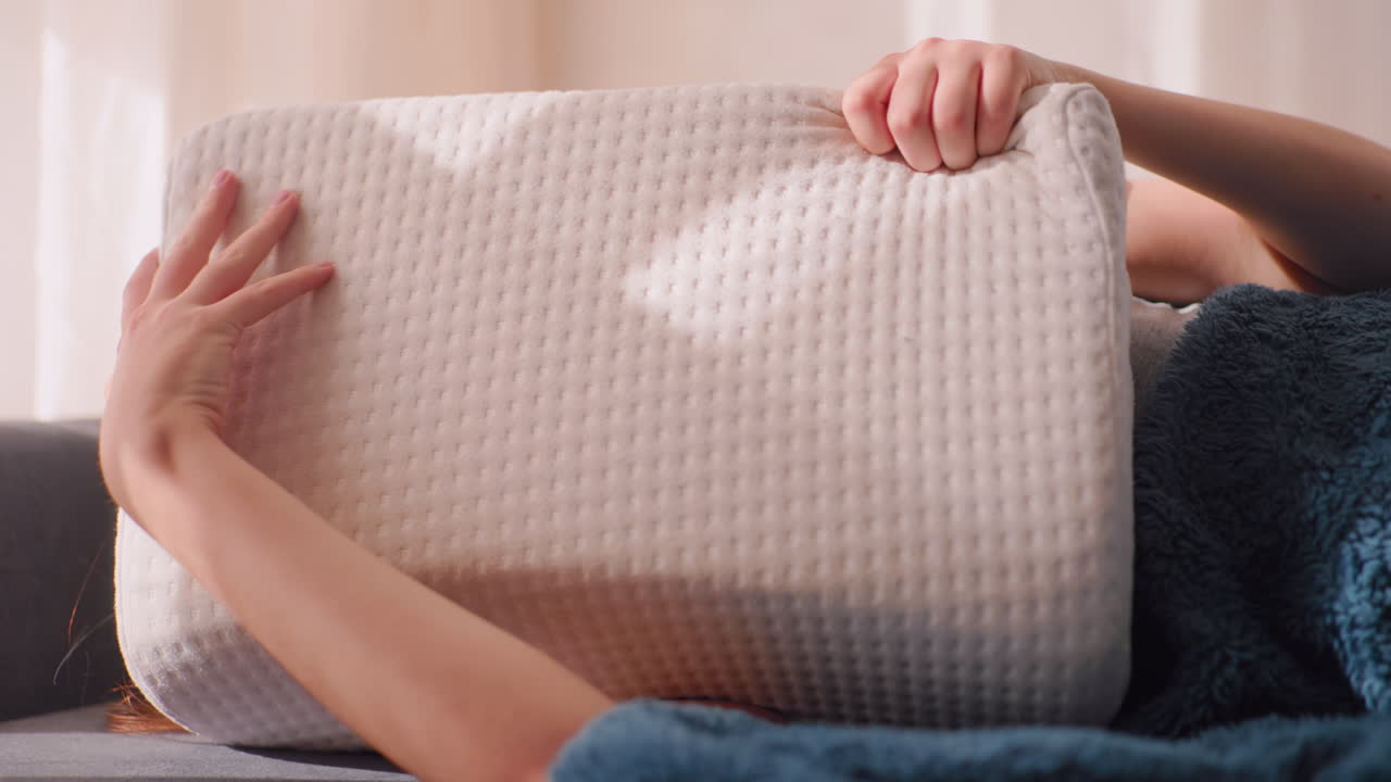 Sleepy woman curled on couch tries to avoid getting up by hiding face under soft pillow, squinting eyes shut against morning light with arm over head, wrapped in fluffy blanket in quiet peaceful room