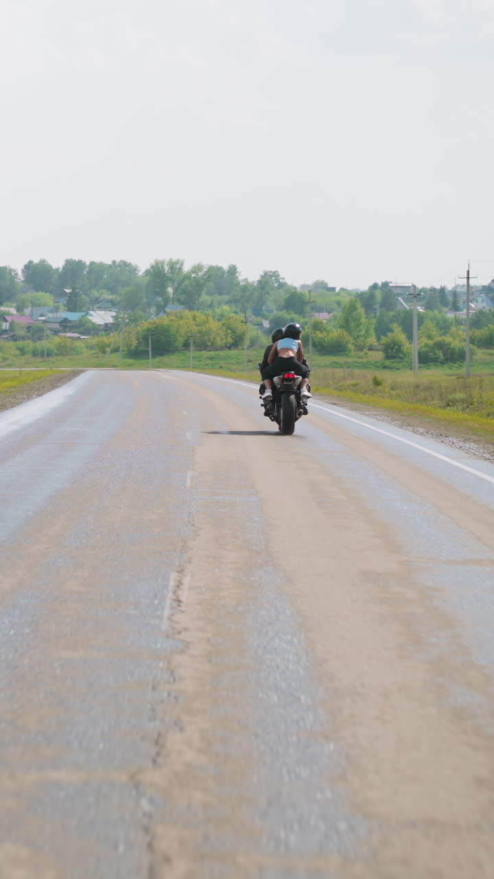 Slim woman with boyfriend biker rides motorcycle on back seat along empty road between green fields at farmland to village backside view slow motion