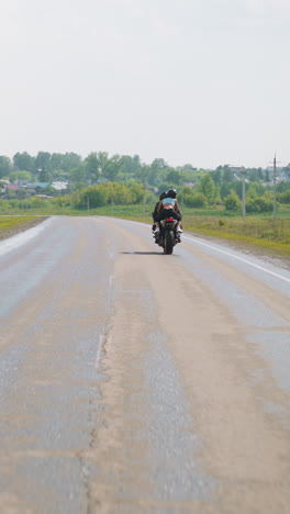 slim woman with boyfriend biker rides motorcycle on back seat along empty road between green fields at farmland to village backside view slow motion