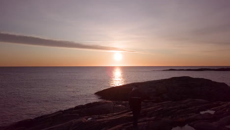 aéreo, disparo de un dron de un hombre caminando en la costa rocosa de la isla de justoya, al amanecer, en aust agder, noruega