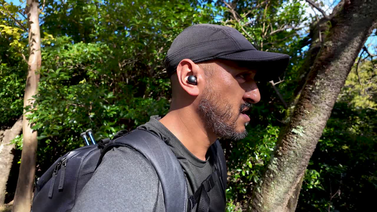A man hikes through a lush forest on a sunny day, wearing earbuds and a backpack, enjoying an outdoor adventure.