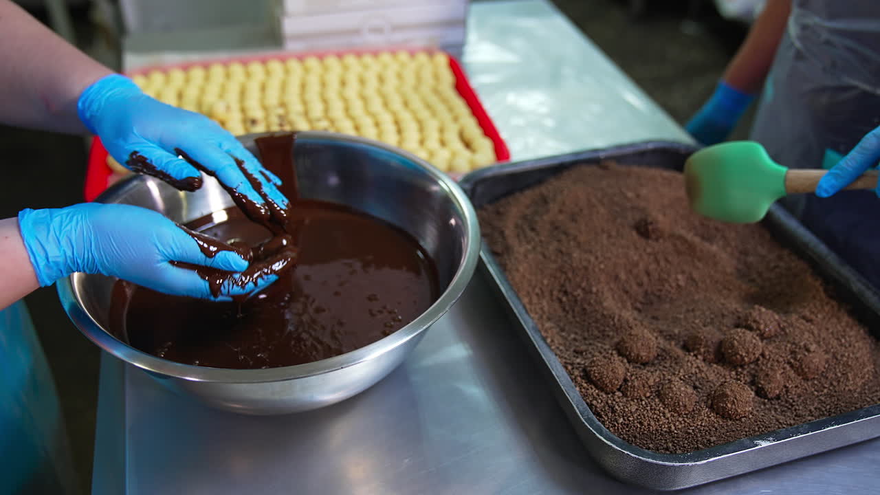 Hands in blue gloves take sweet candy balls and cover them with chocolate. Chocolate sweets are thrown into cocoa and nuts. Other worker mixes candies with sprinkling.