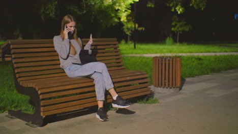 mujer joven sentada en un banco al aire libre por la noche, haciendo una llamada telefónica mientras usa una computadora portátil, iluminada por la luz del parque suave, mochila roja a su lado en el banco, rodeada de vegetación