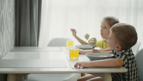 boy and girl sitting at dining table enjoying meal with bowls and glasses of fruit juice in bright room, cheerful kids eating food with large spoons near window with soft natural light