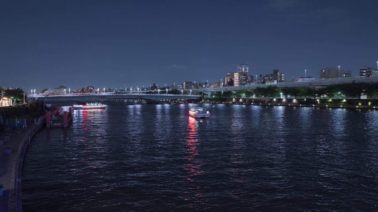 Multiple illuminated boats cruise along Tokyo's vibrant Sumida River at night, creating a bustling scene with city lights reflecting on the water