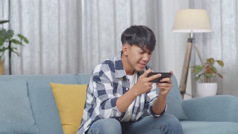 asian teen boy enjoys playing game on smartphone while sitting on sofa in the living room at home