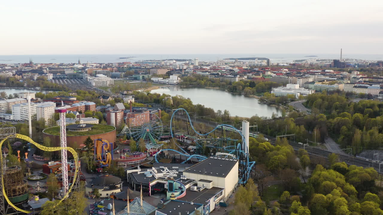 Aerial, pull back, drone shot of a train on the railway, revealing the Linnanmaki amusement park, sunny spring day, in Helsinki, Finland