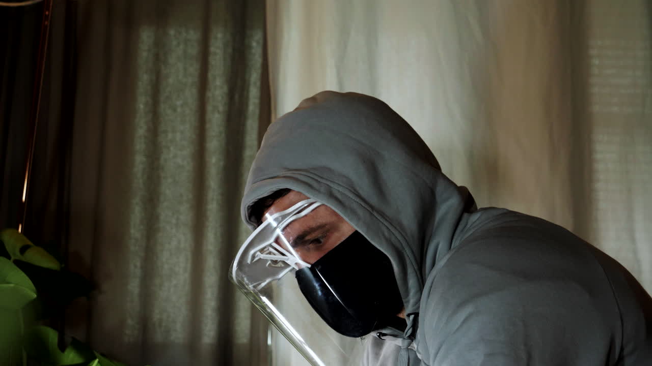 Homem Angustiado usa máscara de proteção e protetor facial sozinho em um quarto escuro em casa