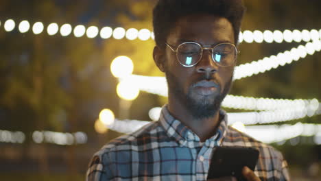 Hombre-Afroamericano-Parado-En-El-Parque-Por-La-Noche-Y-Usando-El-Teléfono