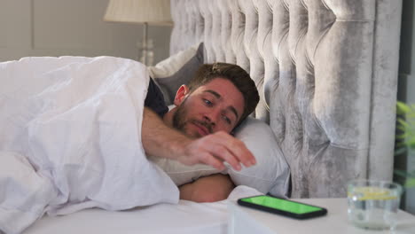 man waking up in bed reaches out to turn off alarm on mobile phone