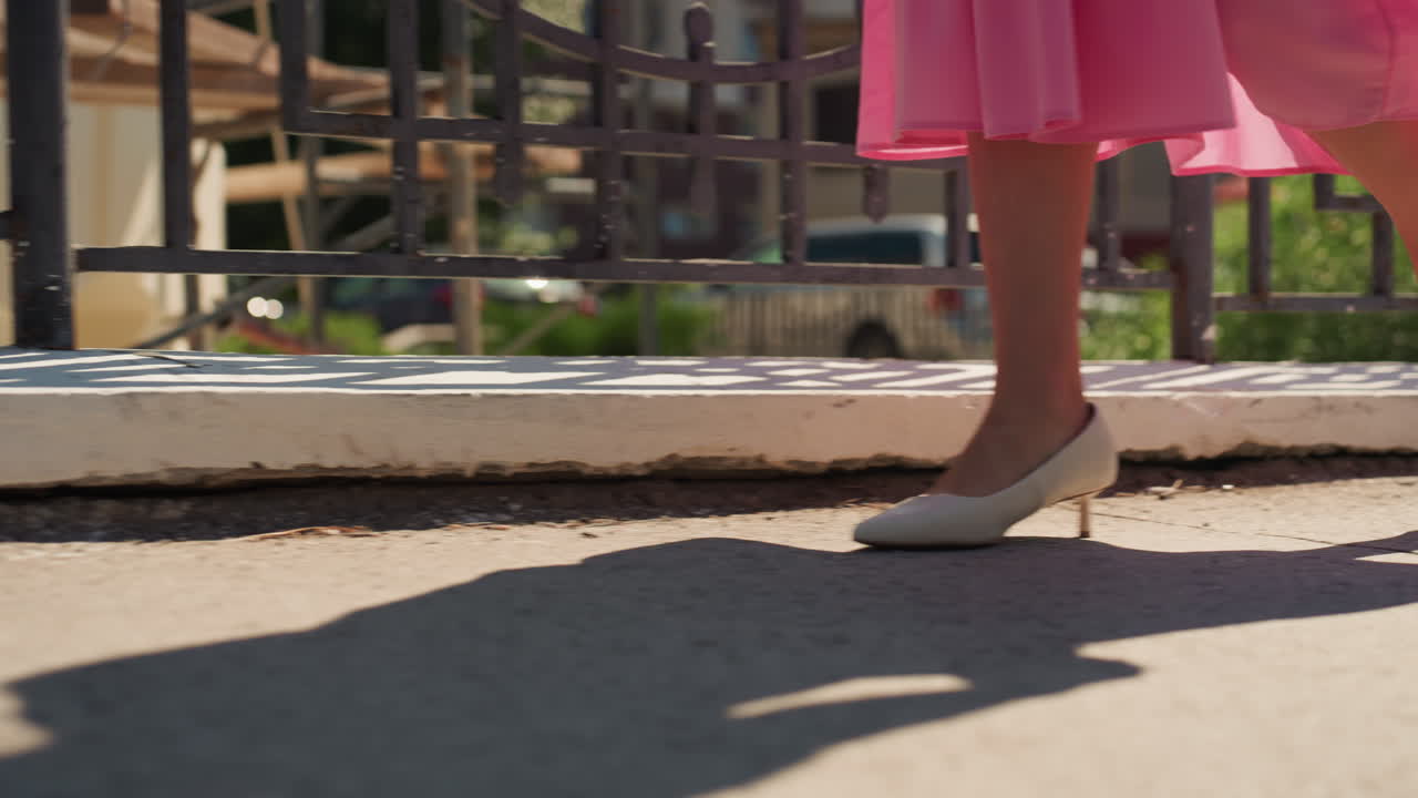 Donna in abito rosa che cammina lungo il marciapiede, primo piano delle gambe e dei tacchi bianchi che proiettano una lunga ombra sul cemento illuminato dal sole vicino a una recinzione decorativa in ferro, atmosfera suburbana delicata, luce pomeridiana soffusa