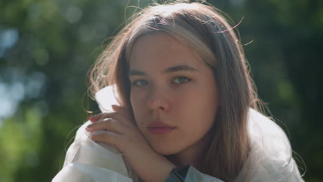 close-up of young woman resting her chin on her hand, with a thoughtful expression and soft sunlight highlighting her face, blurred greenery in background