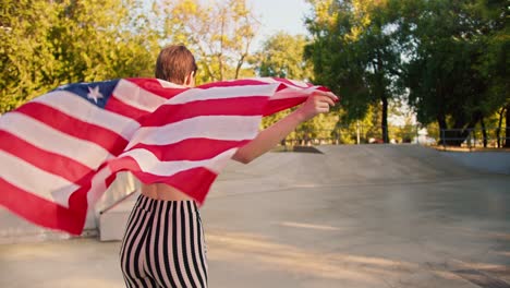 Ein-Glückliches,-Kurzhaariges-Mädchen-In-Einem-Lila-Oberteil-Mit-Roten-Kopfhörern-Fährt-Auf-Rollschuhen-Durch-Einen-Skatepark-Und-Hält-Die-US-Flagge-In-Ihren-Händen